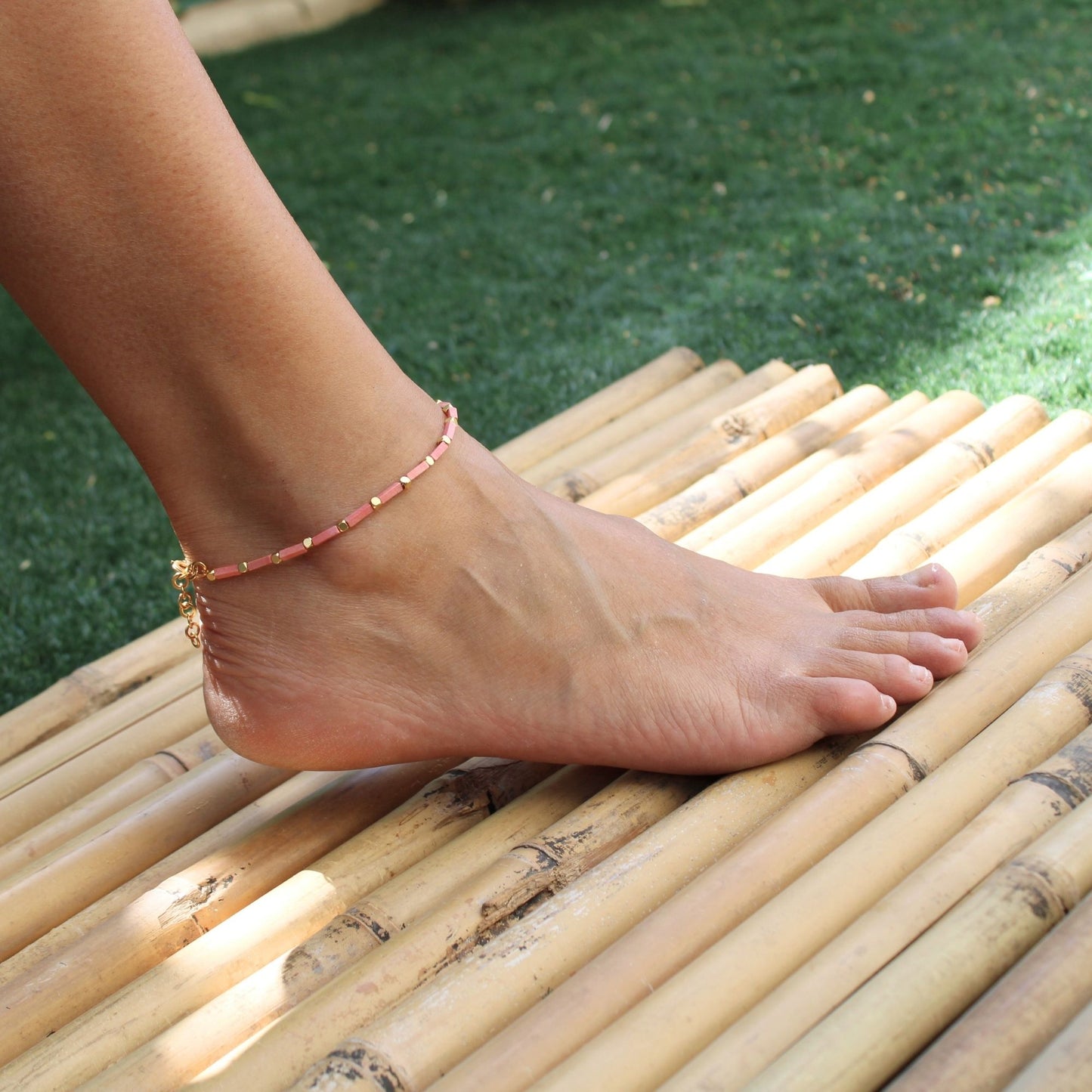 צמיד רגל ורוד עם נגיעות זהב על קרסול נשי, צילום חוץ באור טבעי על משטח במבוק